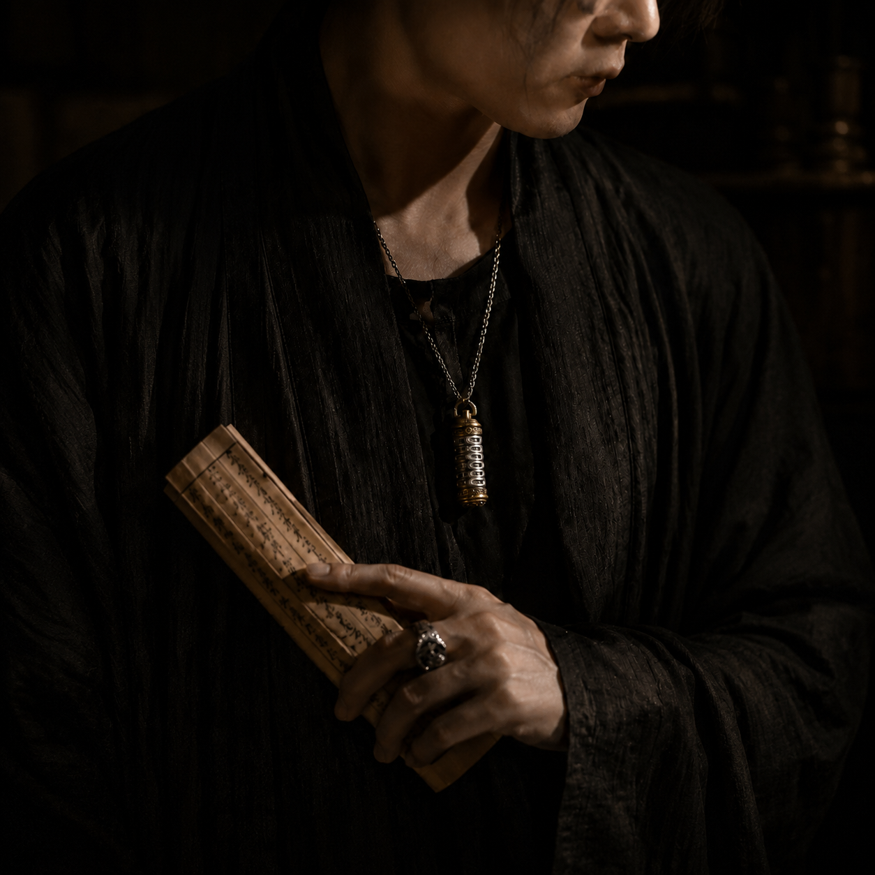Revolving Mantra pendant necklace worn over layered black garments, model holding an antique scroll in a dim atmospheric setting