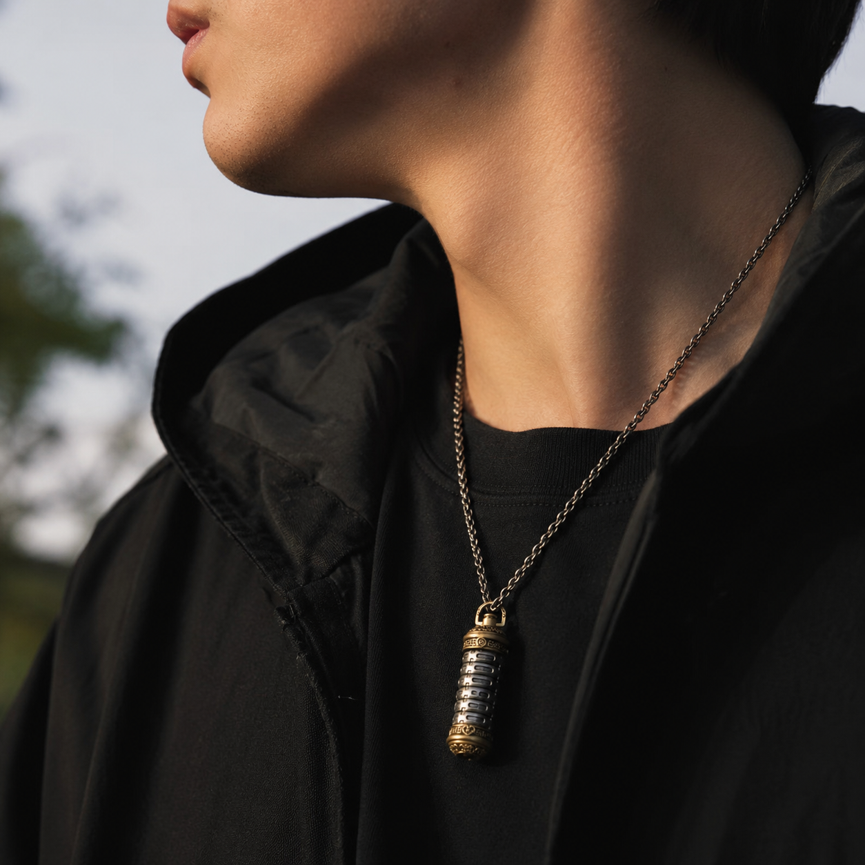 Revolving Mantra pendant necklace worn outdoors over a black hoodie, close-up side angle with natural lighting and blurred foliage background